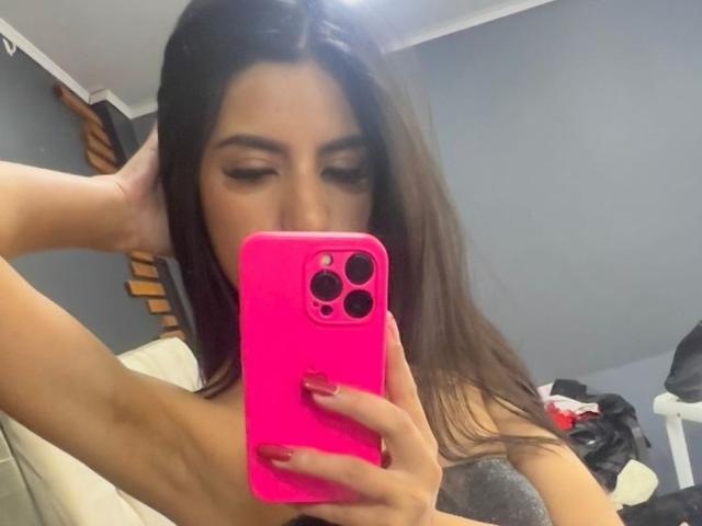 SamiLure, con cabello largo y castaño, sosteniendo un teléfono rosa, tomando una selfie frente al espejo, mostrando su rostro y uñas rojas, en una habitación con paredes grises. Pechos parcialmente visibles, sin ropa..