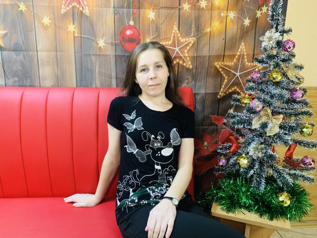 DaisyEdit has medium tits, brown hair, and a serious face, wearing a black shirt with white bird and butterfly designs, sitting on a red couch next to a Christmas tree.