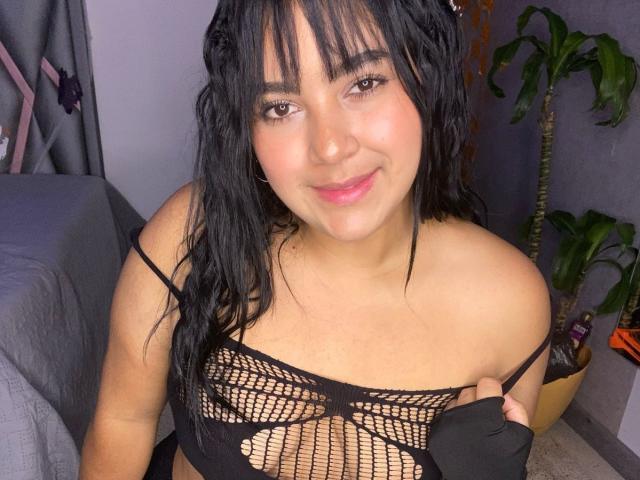 Auroratziu with wet black hair, smiling, wearing a black mesh top that reveals her small tits, in a bedroom with a potted plant and a gray bed.