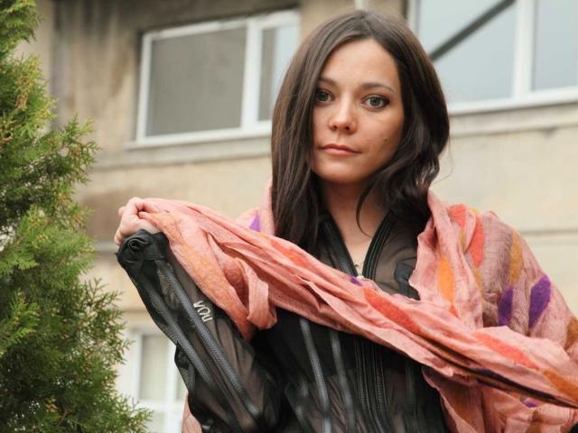 RiaLova with brown hair, wearing a black, zip-up, sheer shirt, holding a pink, semi-transparent, patterned scarf over her shoulders, standing in front of a green tree.