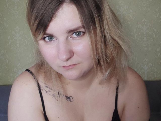 MinistraX has light skin, blue eyes, and blonde-to-brown hair, wearing a black spaghetti strap top with a tattoo on her right shoulder. Her tits are small. She looks serious.