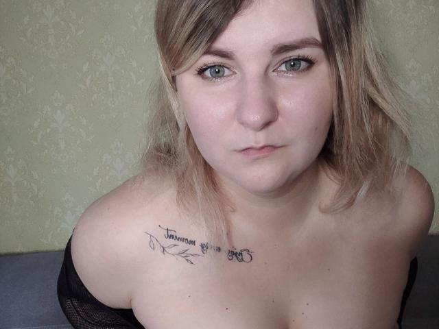 MinistraX has light skin, blue eyes, and blonde shoulder-length hair. She's topless with a tattoo on her right shoulder saying "Tomorrow you will be stronger." Boobs visible.