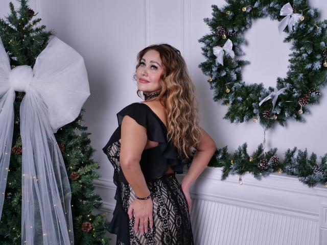 MilaUrban with wavy blonde hair, black lace dress, and choker, standing by Christmas trees, one adorned with a huge white bow, smiling at camera, hand on hip.
