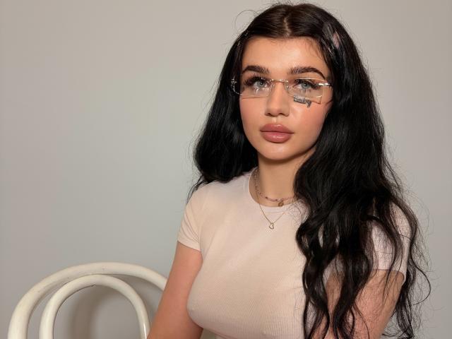 CharlotteFancy with long black hair, wearing glasses and a tight pink top, showing her tits, sitting with a white chair, neutral expression, light skin, subtle makeup, and a necklace.