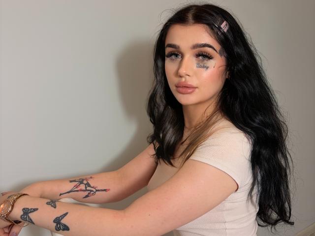CharlotteFancy has long black hair, blue eyes, and wears a beige top. She has tattoos on her arms, including a butterfly and a woman, with a small tattoo on her cheek.