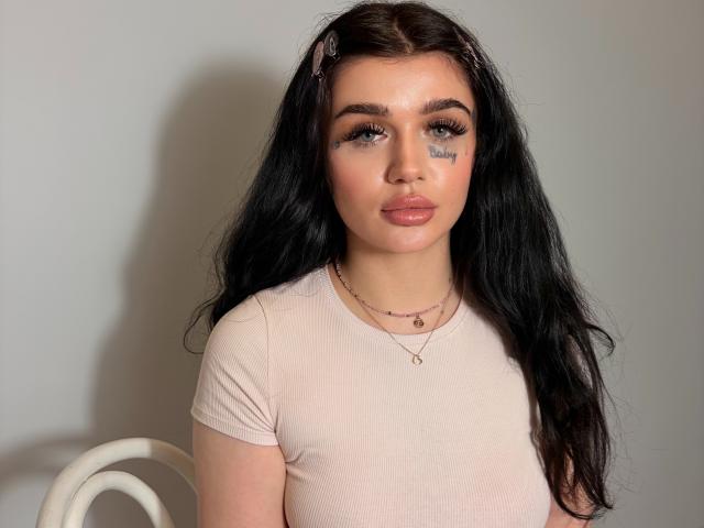 CharlotteFancy has long black hair, full pouty lips, and a tattoo under her right eye. She's wearing a pinkish-beige tee, a pink hairclip, and two necklaces.