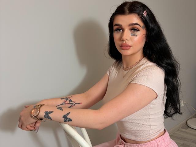 CharlotteFancy with long black hair, pink hairclip, tan shirt, pink pants, tattoos on arms, holding chair, looking at camera, soft makeup, pink lipstick, wrist bracelet, sitting.