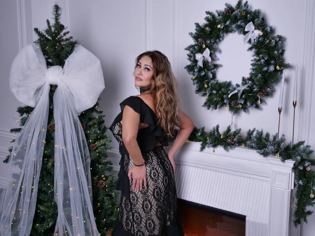 MilaUrban with wavy brown hair, black lace dress, and choker, standing by Christmas tree with white ribbon, and decorated wreath, looking back with hand on hip, showing ass.
