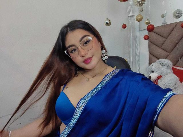 HelueJamil with long brown hair, glasses, blue bra, and matching blue sari, showing her tits, sitting, holding her hair, pink lipstick, and white earrings in a bedroom.