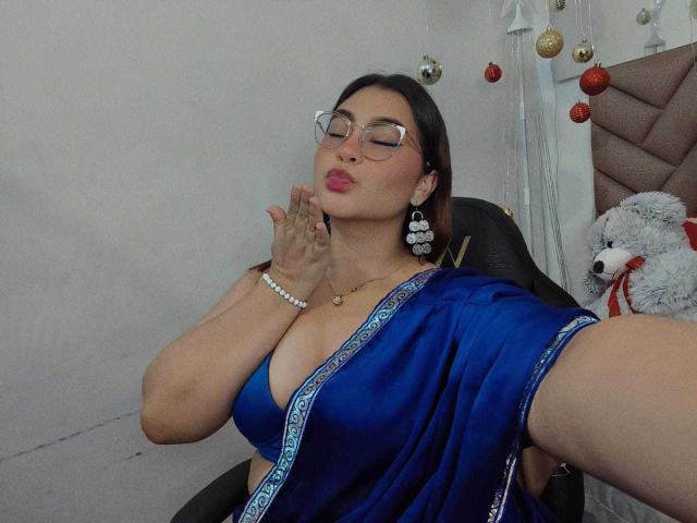 HelueJamil con grandi seni, vestita con un sari blu, con gli occhiali e il rossetto rosa, che bacia la sua mano, seduta su una sedia, indossando un braccialetto di perle e grandi orecchini..