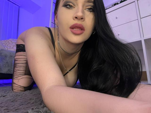 MaryLori, con lunghi capelli neri, indossa una maglietta nera, catene e una manica strappata, è sdraiata su un tappeto grigio, mostrando il seno e un piercing al naso, con illuminazione blu..