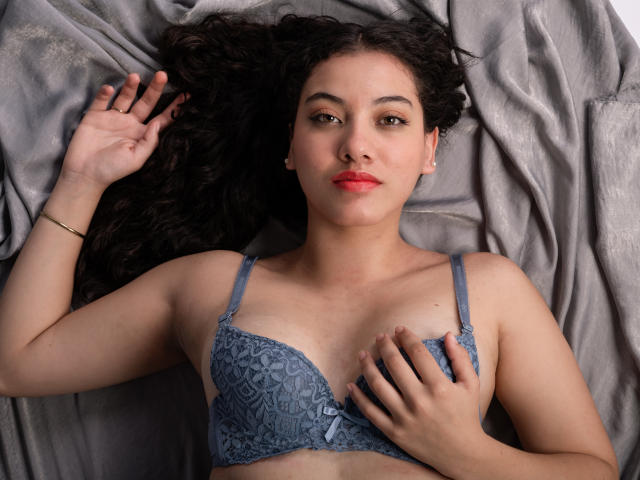 AriannaHills with curly black hair, wearing a blue lace bra, lying on gray fabric, hand on chest, other hand raised, looking at camera, red lipstick, confident expression.