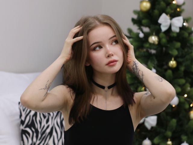 ViolettaFane with hands in brown hair, black sleeveless top, choker, tattoos, and nose ring, looking up with soft pink lips, small breasts, and green eyes. Christmas tree in background.