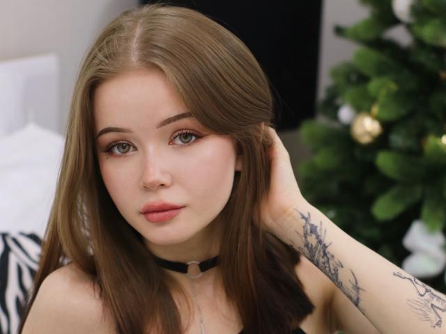 ViolettaFane with long brown hair, green eyes, and pink lips, wearing a black choker, hand in hair, tattoos on right arm, looking at camera with subtle smile.
