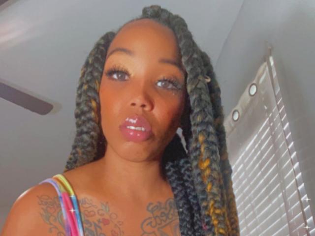 SexxyReddOfficial with long, twisted black and yellow braids, glossy pink lips, and tattoos on her chest, wearing a colorful striped top, looking at the camera with a slight smile.