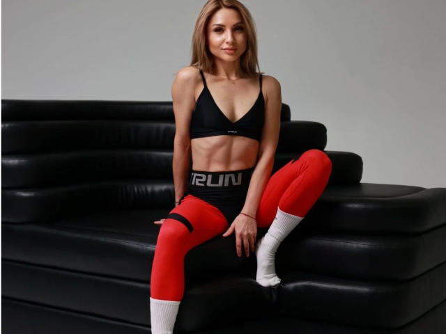 MarynaLove-hot with small tits, wearing a black sports bra, red pants, white socks, sitting on a black leather couch, blonde hair, light skin, and a subtle smile.