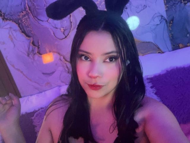 IsabellaCutte has long dark hair, wearing black bunny ears, and a black strapless top. Her makeup is bold with dark eyeliner and red lipstick. Her expression is slightly playful with a subtle smile. The lighting is purple, creating a soft glow on her face and shoulders. Her right hand is partially visible, holding a small object. The texture of the background is fuzzy, with purple and white elements. Her skin is fair, and her eyes are dark.