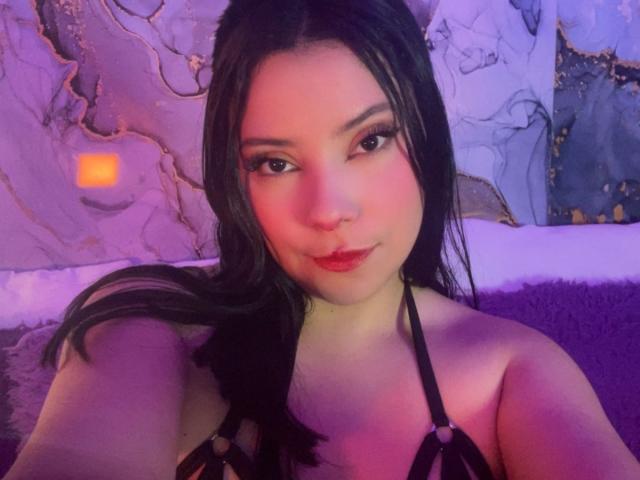 IsabellaCutte with black hair, pink-tinted skin, and red lipstick, wearing a black strappy top, showing her tits, looking at the camera with a slight smile.