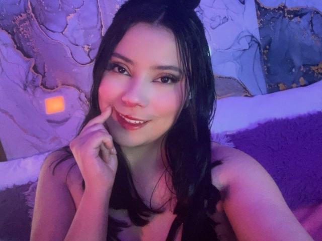 IsabellaCutte with dark hair, smiling, touching her chin, wearing a topless, purple-lit room, her tits partially visible, her finger on her lips, pink lipstick, blue eyes.