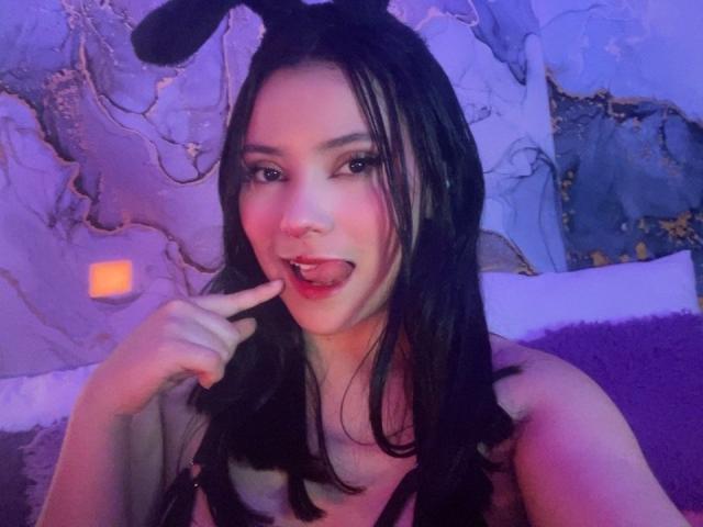 IsabellaCutte with black bunny ears, long dark hair, and a playful expression, sticking her tongue out and touching her lip with her finger, showing her tits.