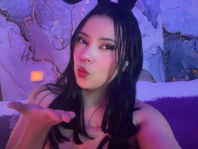 IsabellaCutte with black hair, wearing bunny ears, pouty lips, and making a kissing face, holding up her hand, purple and blue lighting. Her tits are partially visible.