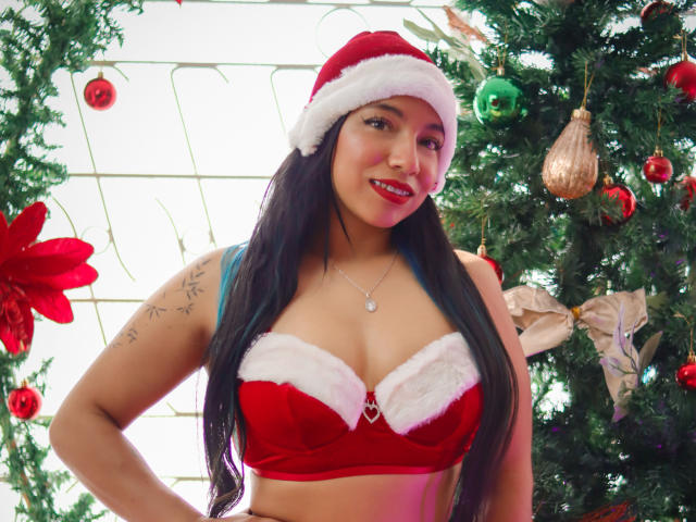 Paulina Colemann, con cabello negro largo, un gorro rojo de Papá Noel, un sujetador rojo de terciopelo con detalles de piel blanca, mostrando sus pechos, una correa azul, un tatuaje en el brazo izquierdo, de pie frente a un árbol de Navidad..