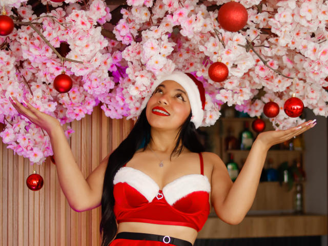 Paulina Colemann, con capelli neri, indossa una maglietta rossa a forma di Babbo Natale in velluto, con bordi di pelliccia bianca, un cappello rosso, sorride, ha le mani alzate ed è circondata da fiori di ciliegio rosa, decorazioni rosse e un muro di legno..
