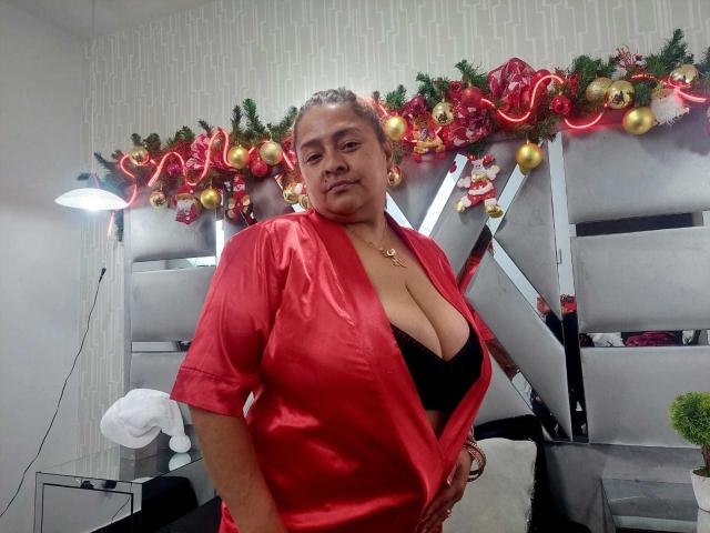 MonzerratBoyer, with large tits, wearing a red satin robe, black bra, and gold necklace, standing in a room with Christmas decorations, looking at the camera, slightly smiling.