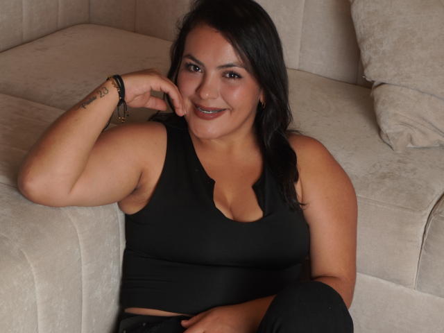 ScarlettOxford, with dark hair and tan skin, sits on a beige sofa in a black tank top, smiling, showing her tits, and resting her chin on her hand.