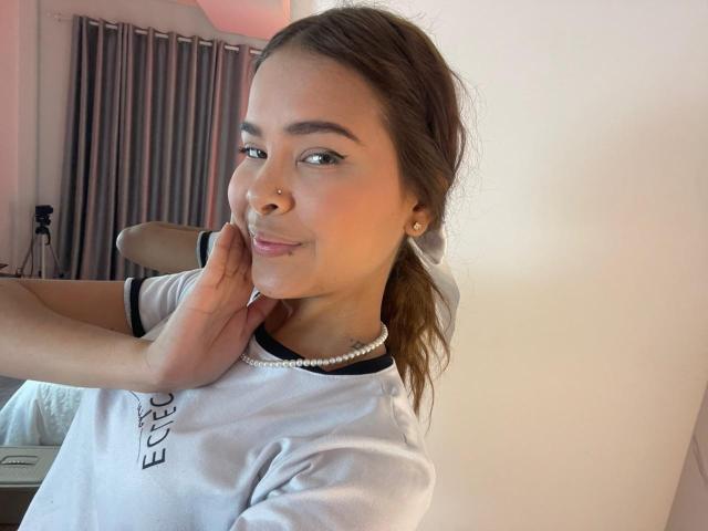ChloeVixen, con un piercing en la nariz, sonriendo, vestida con una camisa blanca con detalles negros, un collar de perlas y cabello castaño recogido en una coleta, tocándose la mejilla. Luz suave y natural..