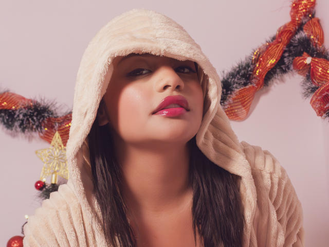 MichelleVain with pink lipstick, wearing a fluffy beige hood, brown hair, and looking up with a slight smirk. Boobs partially visible under the hood. Christmas garland in background.