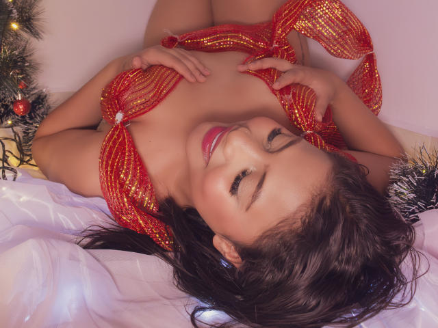 MichelleVain lying on her back with eyes closed, wearing a red sequined dress, hands on her tits, smiling, surrounded by Christmas decorations, and white fabric.