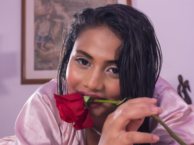 Michelle Vain, avec ses cheveux noirs mouillés, tenant une rose rouge près de ses lèvres légèrement entrouvertes et brillantes, vêtue d'une robe rose en soie, regardant de manière séduisante l'appareil photo..
