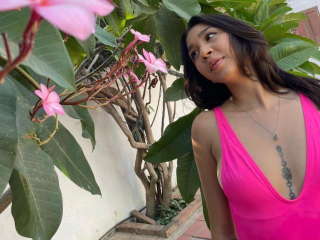 NanaMilan with medium tits in a pink deep V-neck, looking up at pink flowers, has long black hair, a tattoo on her chest, and a necklace, standing by green leaves.