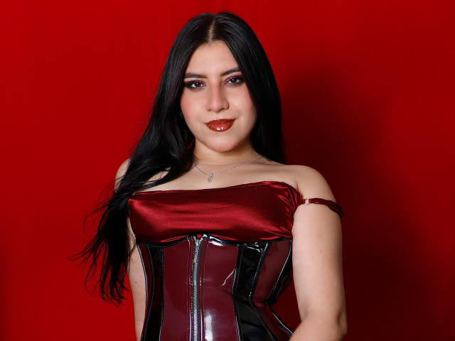 DannaOlivieri with long black hair, red lipstick, and a shiny red and black leather corset, smiling at the camera. Boobs are not visible. Her pussy is not shown.