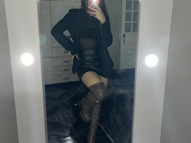 Mirrored selfie of SofiaPassionne. She's wearing a black, sheer mesh crop top, a short black leather skirt, and a black blazer. Her left hand is on her hip, and she's lifting her right leg, showing brown knee-high boots. Her curly hair is partially visible. The reflection shows two bright lights on either side of the mirror. The room has a white door with a decorative design and a white dresser in the background. The floor is dark with a pattern.