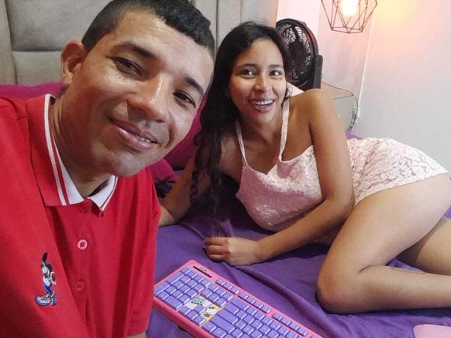 JazminaAndJosephe in pink lace lingerie, lying on purple bed, smiling, with Josephe in red polo, short hair, holding pink keyboard, bedroom, both looking at camera, Josephe on left.