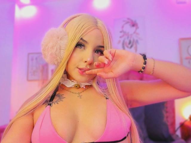 GraceClarke with long blonde hair, pink bikini top, fluffy white earphones, choker, tattoo on left chest, touching nose, pink and purple lights, smiling, bracelet on right wrist.