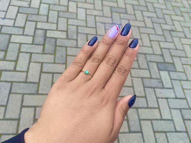 La mano de SophieSonyas, con esmalte de uñas azul oscuro, una uña a rayas moradas y blancas, y un anillo verde con una pequeña piedra azul, sobre un patrón de ladrillos grises..