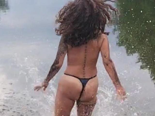 SamsonLili è senza maglietta, indossa uno slip nero, è in piedi nell'acqua, con la schiena rivolta verso la telecamera, mostrando un tatuaggio lungo la colonna vertebrale, con capelli castani ricci e un braccio tatuato..