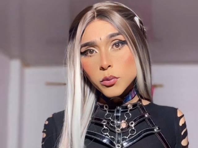 Zasha69 has long, straight, white-blonde hair with dark roots, wearing a black leather top with metal chains, and has a cute, pouty face with brown eyes and pink lips.