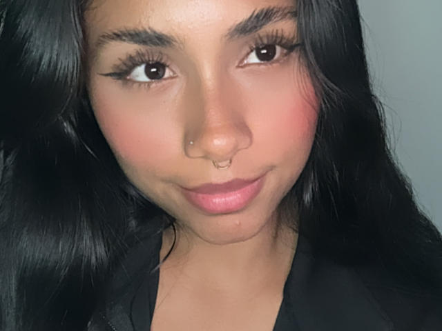 AnaRome with black hair, brown eyes, and a nose ring, smiling softly, wearing a black top, showing pink lips and a slight blush on her face.