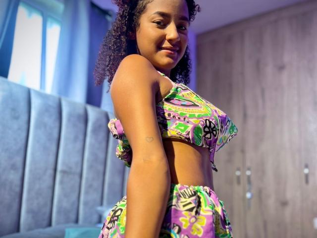 LeilaZahar with curly hair, medium brown skin, small tits, wearing colorful, patterned bikini top and bottom, smiling slightly, standing in bedroom with blue upholstered bed.