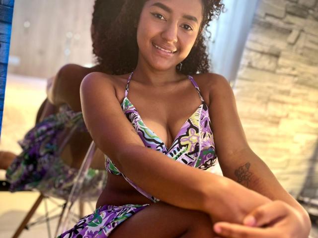 LeilaZahar, dark-skinned woman with curly hair, smiling, wearing a colorful patterned bikini, showing her small boobs, tattoo on right forearm, sitting with arms crossed, indoor setting.