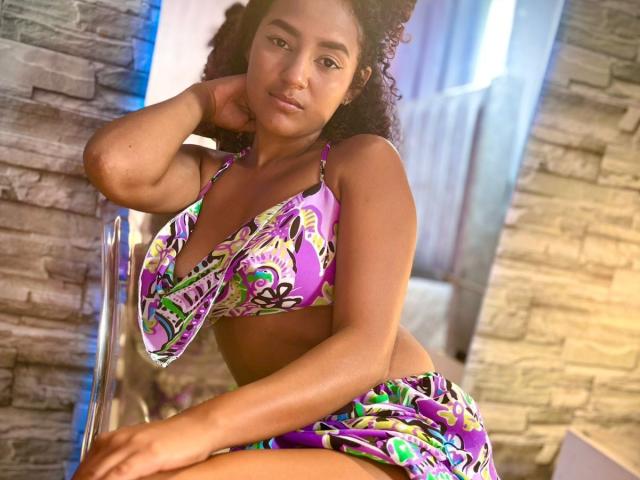 LeilaZahar with curly brown hair, medium brown skin, wearing a purple and green patterned bikini, sitting on a chair, her right hand behind her head, looking at the camera.