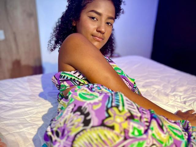 LeilaZahar with curly hair, brown skin, and a pretty face, sits on a white bed, wearing a colorful, patterned dress, covering her ass, looking at the camera.