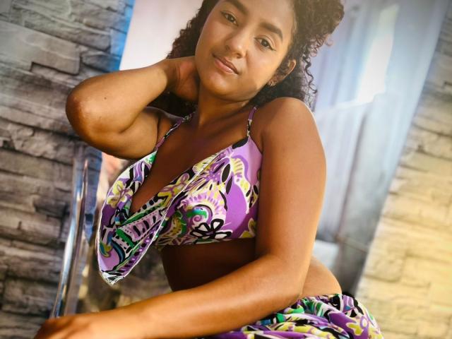 LeilaZahar with curly brown hair, medium brown skin, wearing a purple and green patterned bikini top, smiling, hand behind head, showing small tits and toned belly.