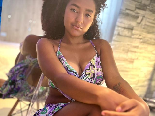 LeilaZahar with curly hair, wearing a purple and green floral bikini, showing her small tits and tattoo on her right arm, sitting with a relaxed, confident expression.