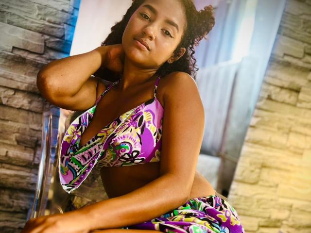 LeilaZahar with curly dark hair, brown skin, wearing a colorful, patterned bikini top and bottom, leaning on a chair, looking at the camera with a slight smile.