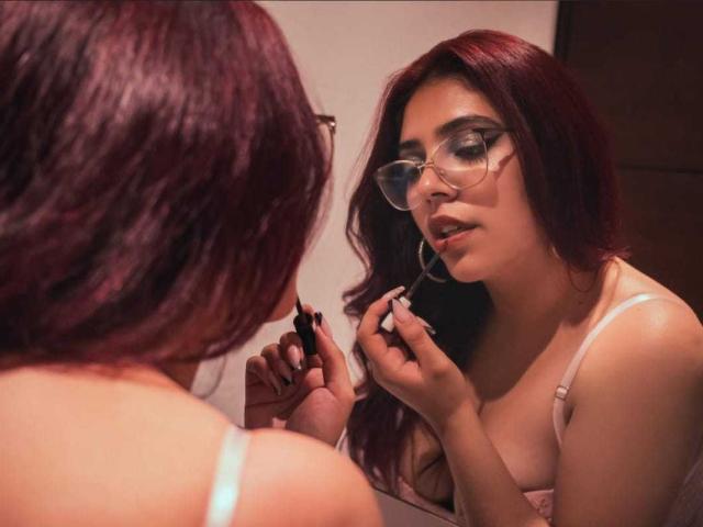 RousseColliinss with red hair, glasses, and pink bra, applying lipstick in a mirror, showing her tits and holding a makeup brush. Reflection shows her focused face.